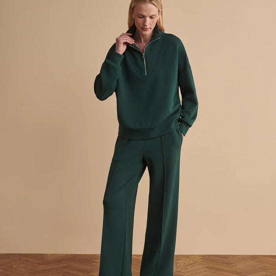 A woman modeling a dark green two-piece tracksuit consisting of a quarter-zip long-sleeve top and matching wide-leg trousers, paired with white sneakers, set against a beige studio background with a wood herringbone floor.