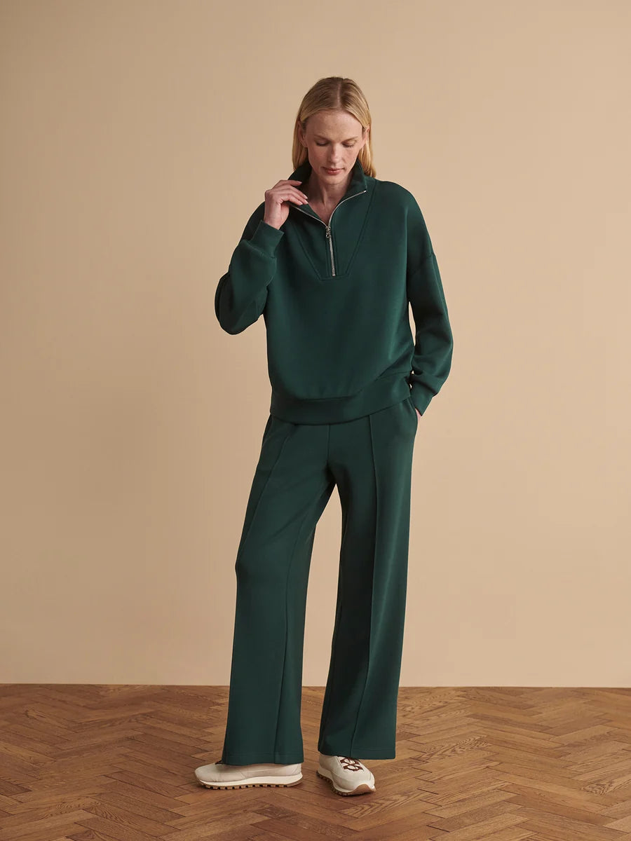 A woman modeling a dark green two-piece tracksuit consisting of a quarter-zip long-sleeve top and matching wide-leg trousers, paired with white sneakers, set against a beige studio background with a wood herringbone floor.
