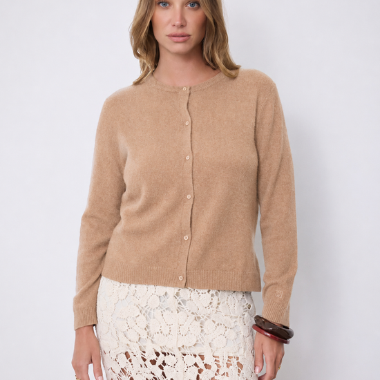 A woman shown wearing a cropped, long-sleeve knit cardigan in a warm camel tan color over a white lace skirt, set against a plain light gray studio background.