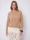 A woman shown wearing a cropped, long-sleeve knit cardigan in a warm camel tan color over a white lace skirt, set against a plain light gray studio background.