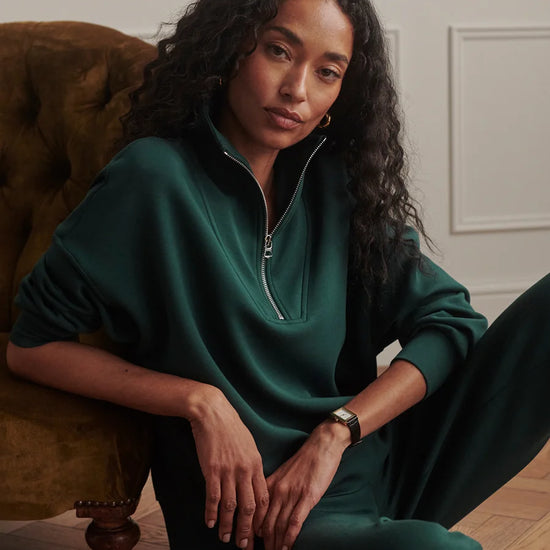 A woman with long curly dark hair sits on a brown velvet armchair wearing a dark green half-zip sweatshirt and matching green pants in a styled indoor setting.
