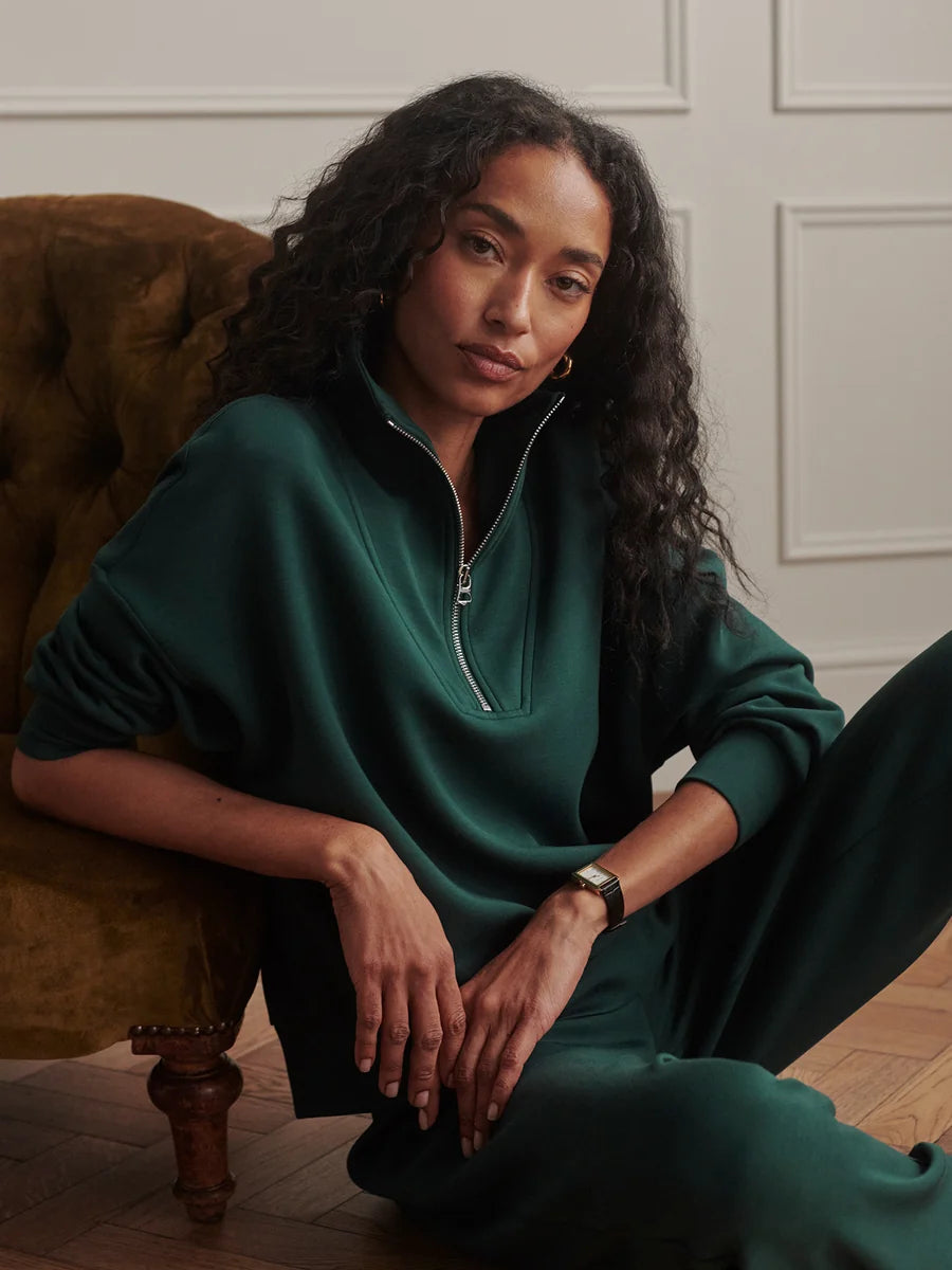 A woman with long curly dark hair sits on a brown velvet armchair wearing a dark green half-zip sweatshirt and matching green pants in a styled indoor setting.