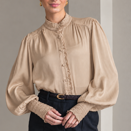 A woman wearing a beige long-sleeve blouse with a high gathered mock neck, decorative lace trim down the front, and voluminous balloon sleeves with textured lace cuffs, paired with dark navy trousers.