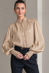 A woman wearing a beige long-sleeve blouse with a high gathered mock neck, decorative lace trim down the front, and voluminous balloon sleeves with textured lace cuffs, paired with dark navy trousers.