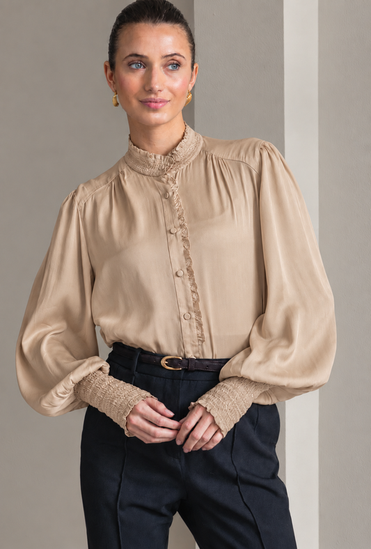 A woman wearing a beige long-sleeve blouse with a high gathered mock neck, decorative lace trim down the front, and voluminous balloon sleeves with textured lace cuffs, paired with dark navy trousers.