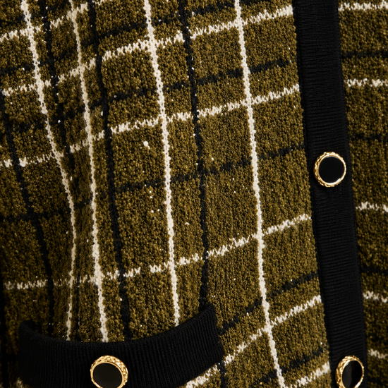 Close-up of a textured knit cardigan featuring an olive green, black, and cream plaid pattern, with a black ribbed trim and gold-tone metal grommets.