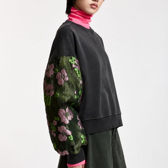 A model wearing a black crewneck sweatshirt with sheer green floral-patterned puff sleeves, layered over a bright pink turtleneck and paired with a dark green skirt, against a plain light gray background.