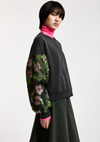 A model wearing a black crewneck sweatshirt with sheer green floral-patterned puff sleeves, layered over a bright pink turtleneck and paired with a dark green skirt, against a plain light gray background.