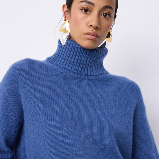 A woman wearing a chunky knit turtleneck sweater in a medium periwinkle blue color, paired with large gold geometric earrings, against a plain light gray background.