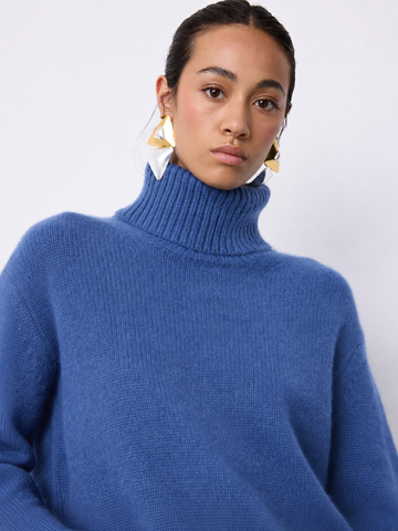 A woman wearing a chunky knit turtleneck sweater in a medium periwinkle blue color, paired with large gold geometric earrings, against a plain light gray background.