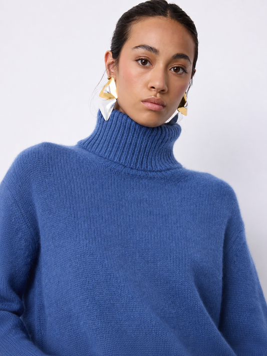 A woman wearing a chunky knit turtleneck sweater in a medium periwinkle blue color, paired with large gold geometric earrings, against a plain light gray background.