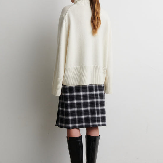 Person wearing a white turtleneck, plaid skirt, and black rain boots against a white wall from Second Female