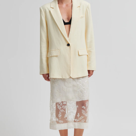 Woman wearing a light yellow blazer over a black bralette and floral skirt on a white background