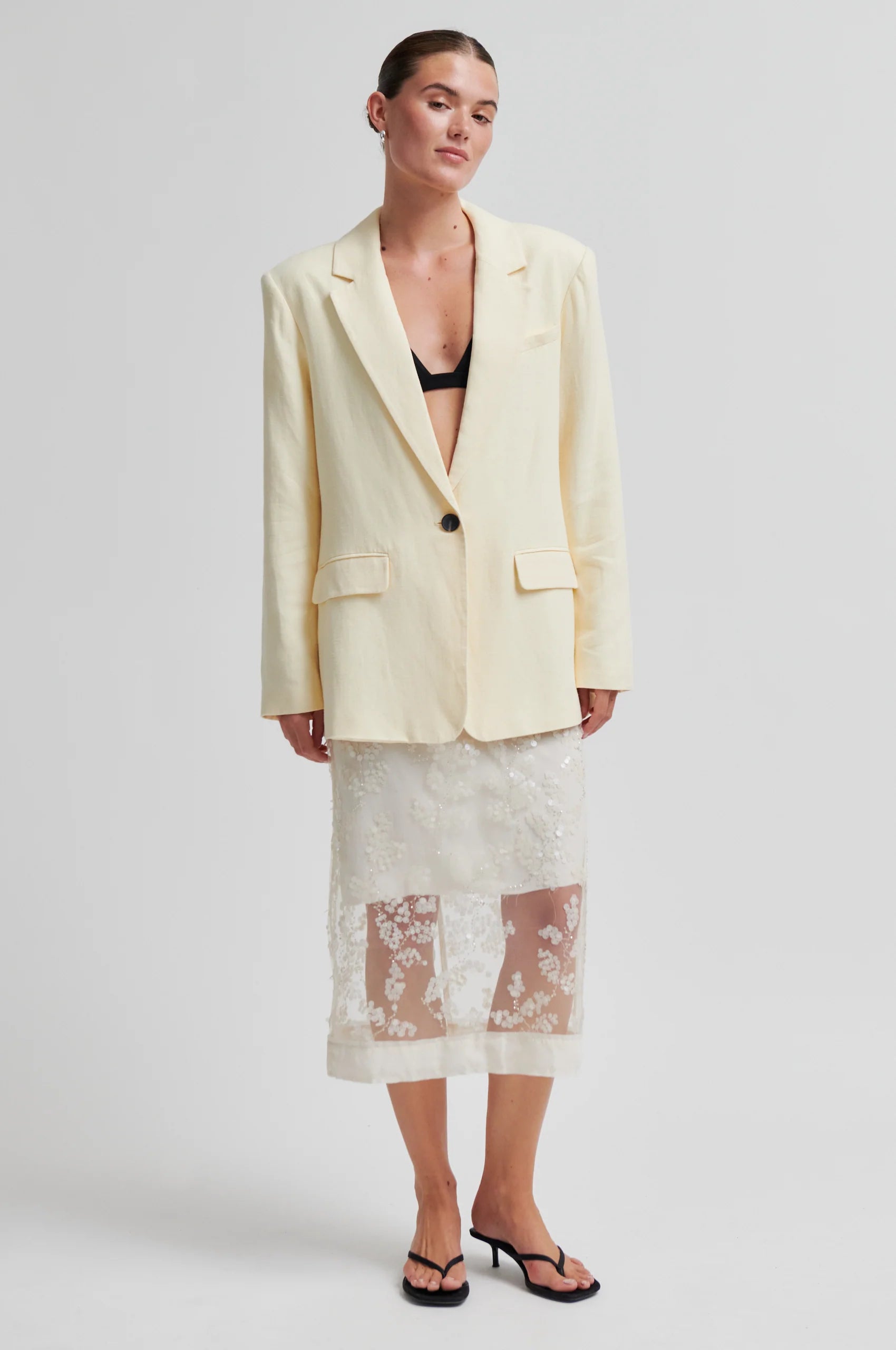 Woman wearing a light yellow blazer over a black bralette and floral skirt on a white background