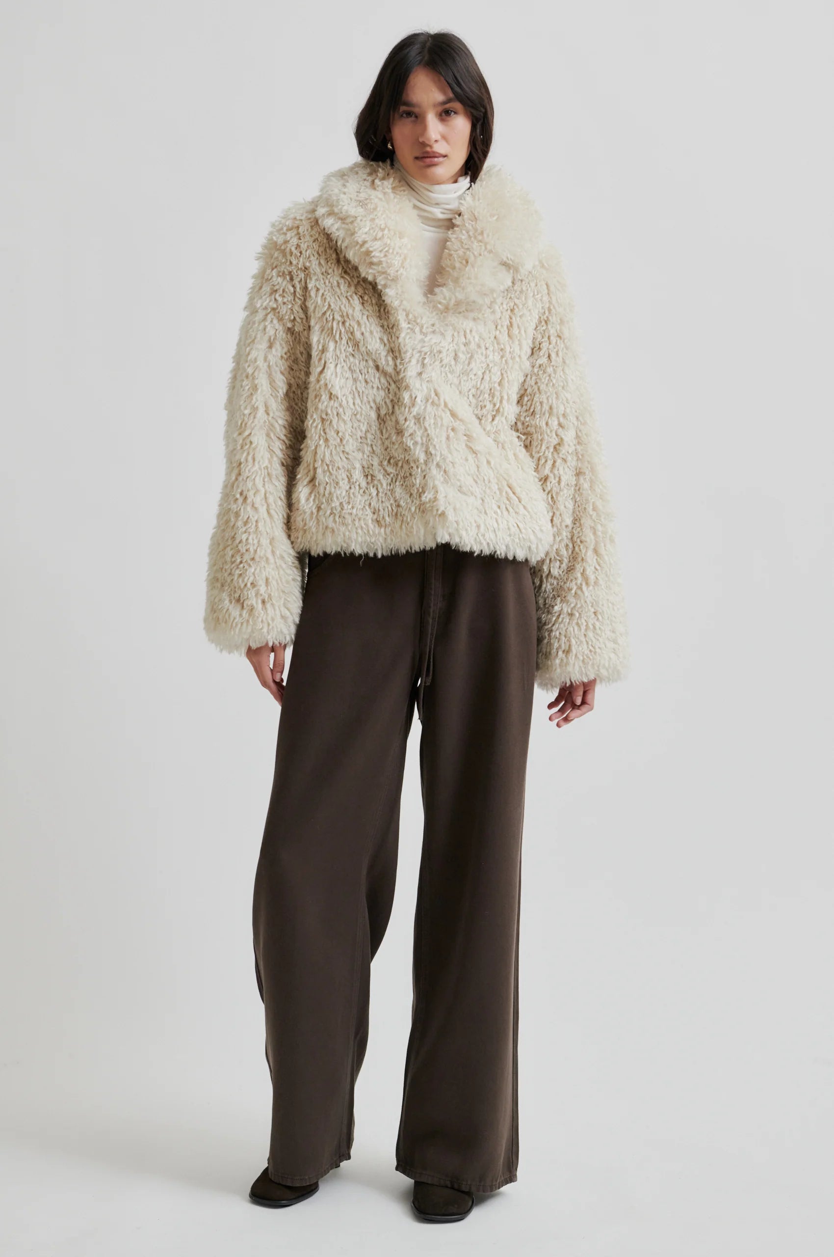 Person wearing a fluffy beige coat and dark pants on a white background