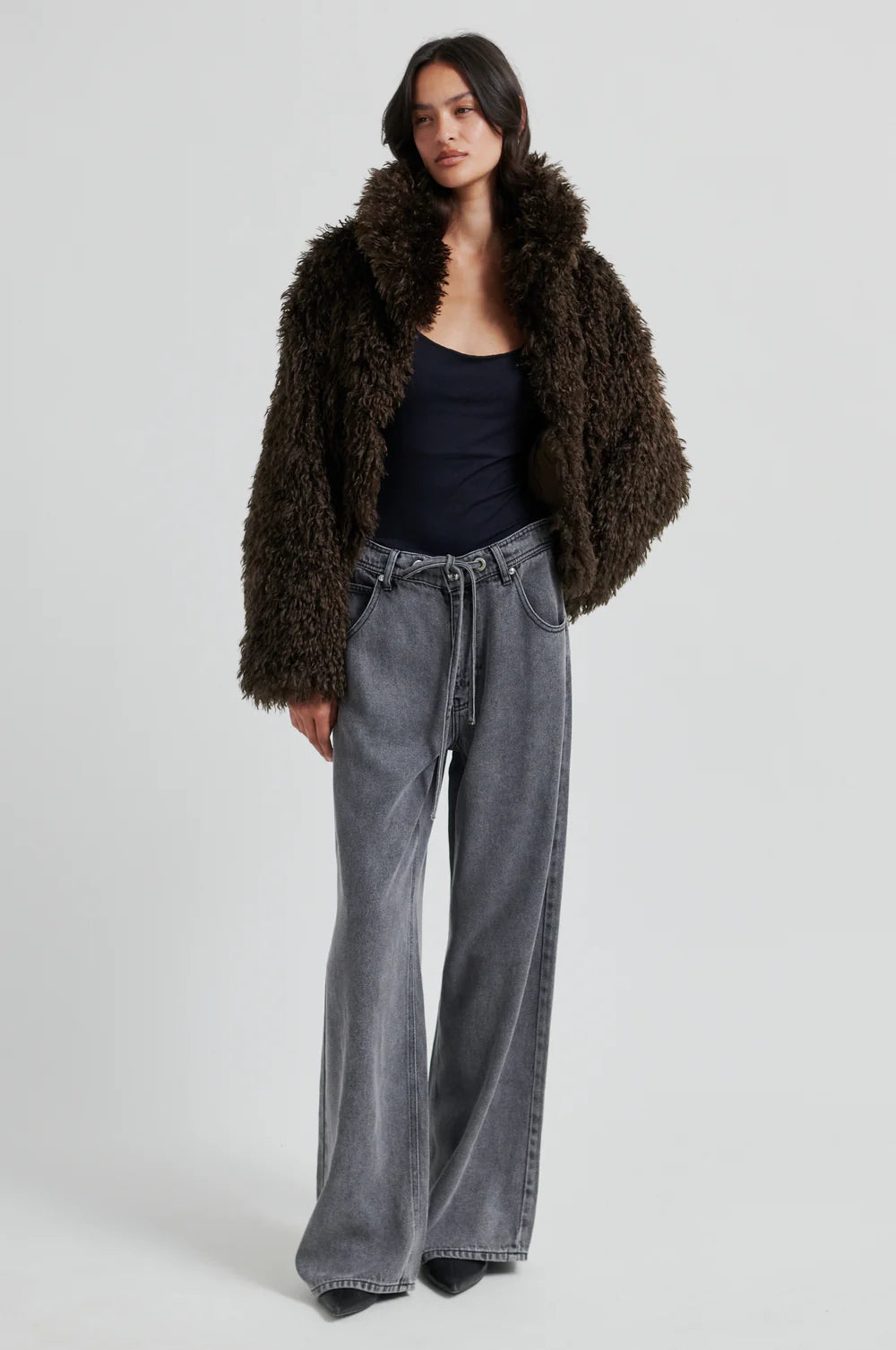 Woman wearing a brown fur coat, black top, and gray jeans on a white background