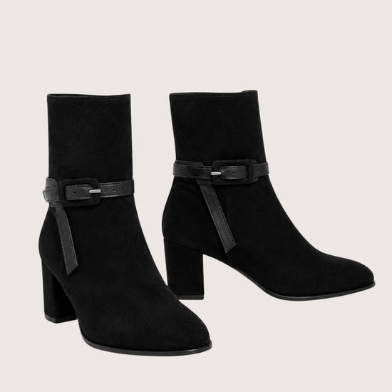 A pair of black suede ankle boots with block heels and a decorative strap detail across the ankle, shown against a light beige background.