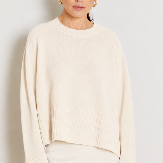 A woman wearing a cream-colored knit sweater with a crew neck and flared cuffs, paired with a matching cream skirt, set against a plain light gray background.