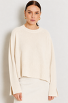 A woman wearing a cream-colored knit sweater with a crew neck and flared cuffs, paired with a matching cream skirt, set against a plain light gray background.