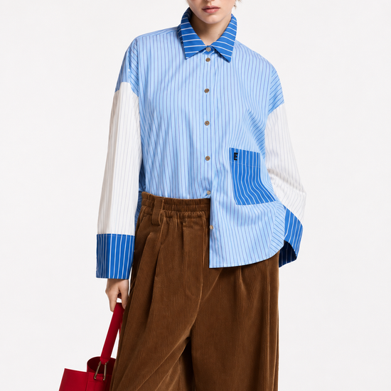 A model wearing a light blue and white color-blocked long-sleeve shirt with blue striped details, paired with wide-leg brown corduroy trousers, black sunglasses, and black shoes, holding a red and blue bag against a plain white studio background.