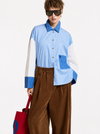 A model wearing a light blue and white color-blocked long-sleeve shirt with blue striped details, paired with wide-leg brown corduroy trousers, black sunglasses, and black shoes, holding a red and blue bag against a plain white studio background.