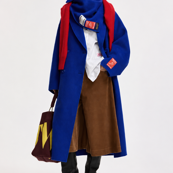 A model stands against a light gray studio background wearing a long blue coat over a white shirt and brown skirt, with a bright red scarf and black knee-high boots. A large brown tote bag with yellow lettering is held in one hand.