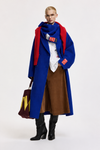 A model stands against a light gray studio background wearing a long blue coat over a white shirt and brown skirt, with a bright red scarf and black knee-high boots. A large brown tote bag with yellow lettering is held in one hand.