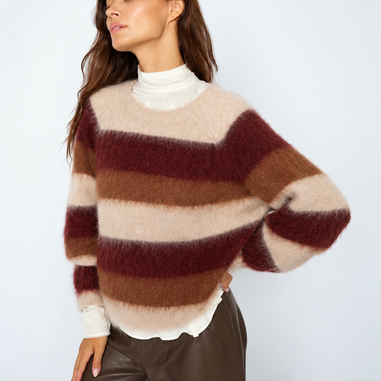 A woman wearing a fuzzy, horizontally striped knit sweater in cream, brown, and burgundy, layered over a white mock neck top and paired with dark brown leather pants, set against a plain light gray studio background.