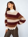 A woman wearing a fuzzy, horizontally striped knit sweater in cream, brown, and burgundy, layered over a white mock neck top and paired with dark brown leather pants, set against a plain light gray studio background.