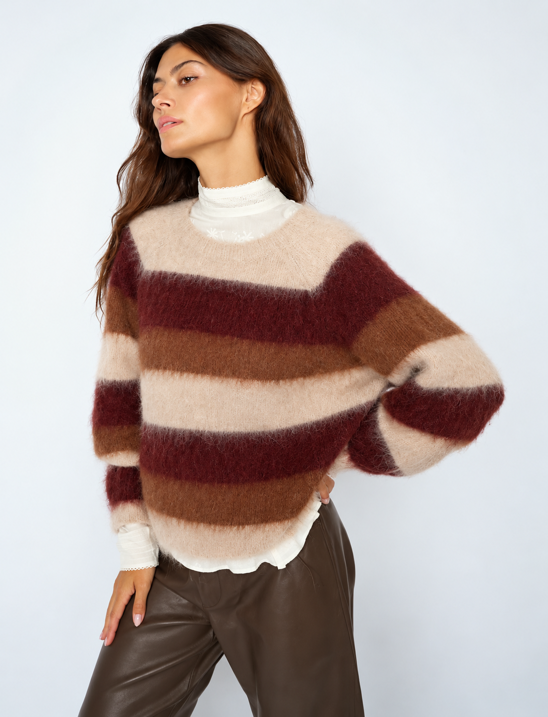 A woman wearing a fuzzy, horizontally striped knit sweater in cream, brown, and burgundy, layered over a white mock neck top and paired with dark brown leather pants, set against a plain light gray studio background.