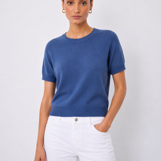 A woman modeling a short-sleeve crewneck knit sweater in a medium blue color, paired with white pants, against a light gray studio background.