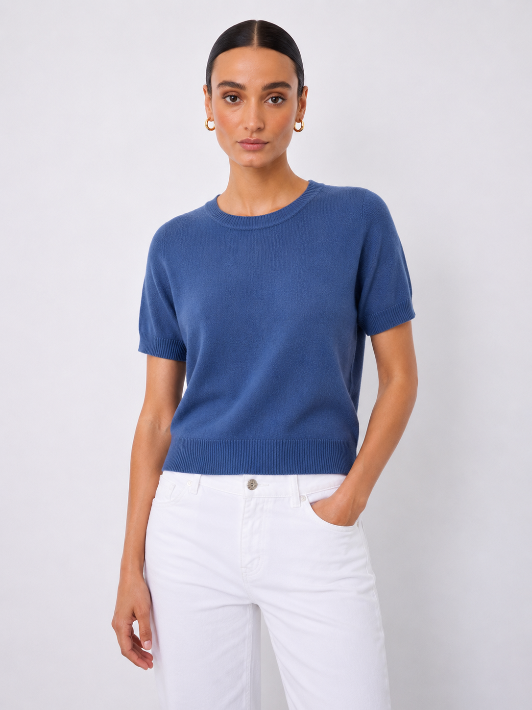 A woman modeling a short-sleeve crewneck knit sweater in a medium blue color, paired with white pants, against a light gray studio background.
