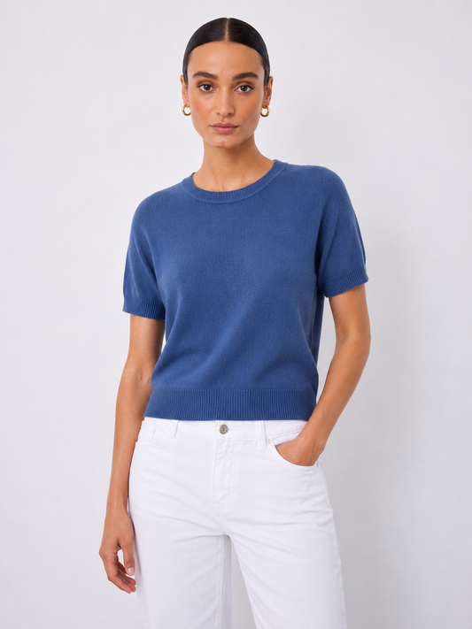 A woman modeling a short-sleeve crewneck knit sweater in a medium blue color, paired with white pants, against a light gray studio background.
