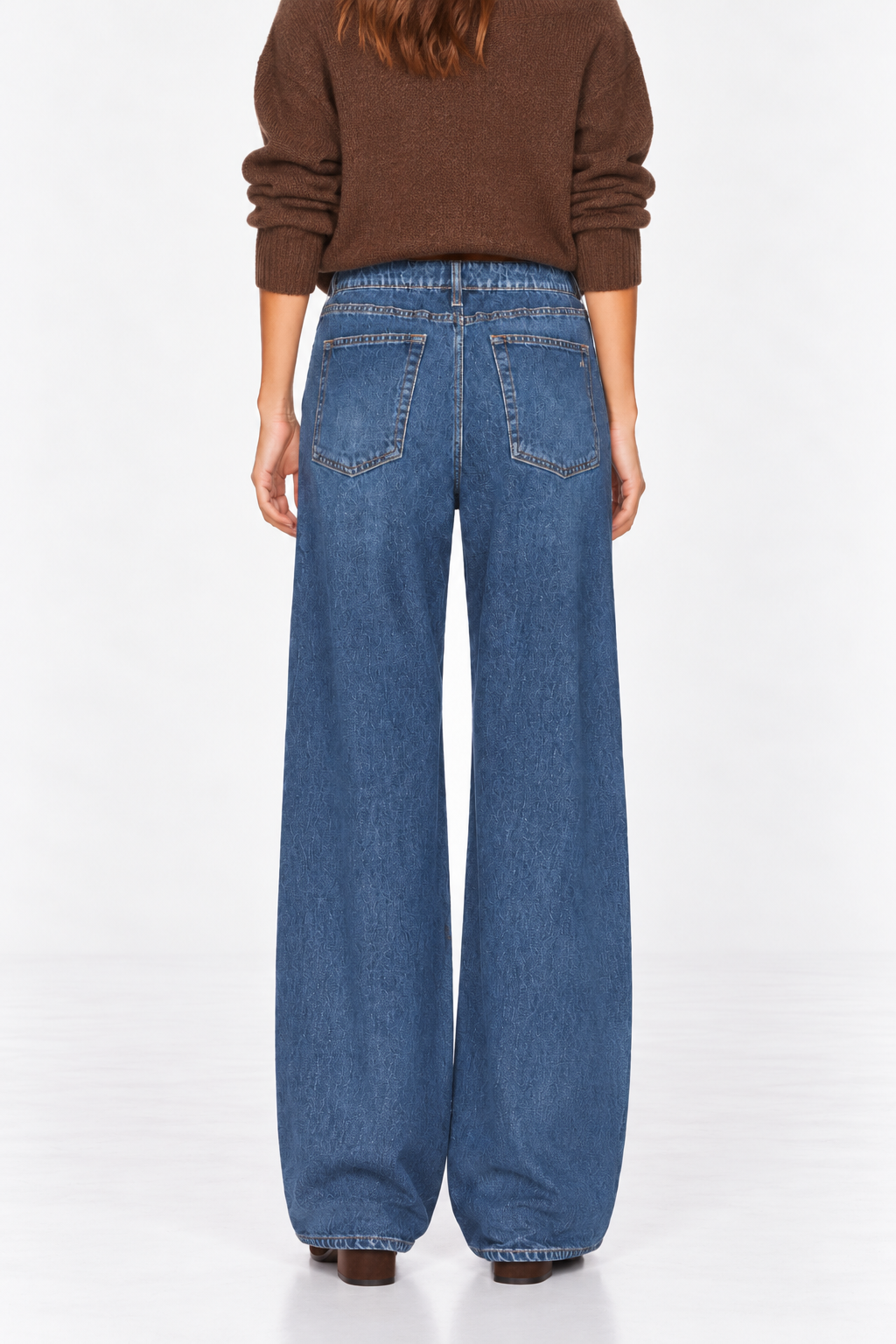 A person shown from the back wearing wide-leg blue denim jeans and a brown long-sleeve top against a plain white studio background.