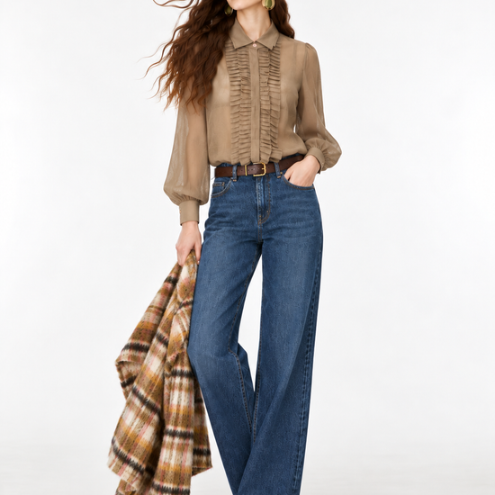 A woman modeling a beige long-sleeve blouse with vertical ruffles, paired with blue wide-leg jeans and holding a brown and beige plaid coat against a plain white studio background.