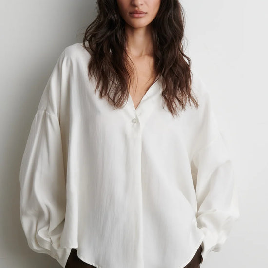 A woman wearing a loose-fitting, off-white button-up blouse with voluminous balloon sleeves and dark brown trousers, standing against a plain light gray wall.