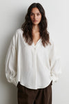 A woman wearing a loose-fitting, off-white button-up blouse with voluminous balloon sleeves and dark brown trousers, standing against a plain light gray wall.