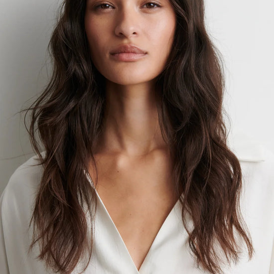 A woman with long, wavy dark brown hair wearing a white button-down shirt, shown from the chest up against a plain light gray background.
