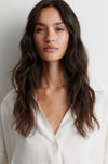 A woman with long, wavy dark brown hair wearing a white button-down shirt, shown from the chest up against a plain light gray background.
