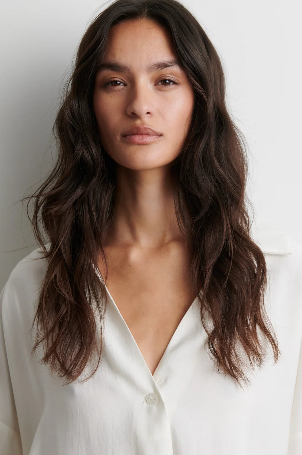 A woman with long, wavy dark brown hair wearing a white button-down shirt, shown from the chest up against a plain light gray background.