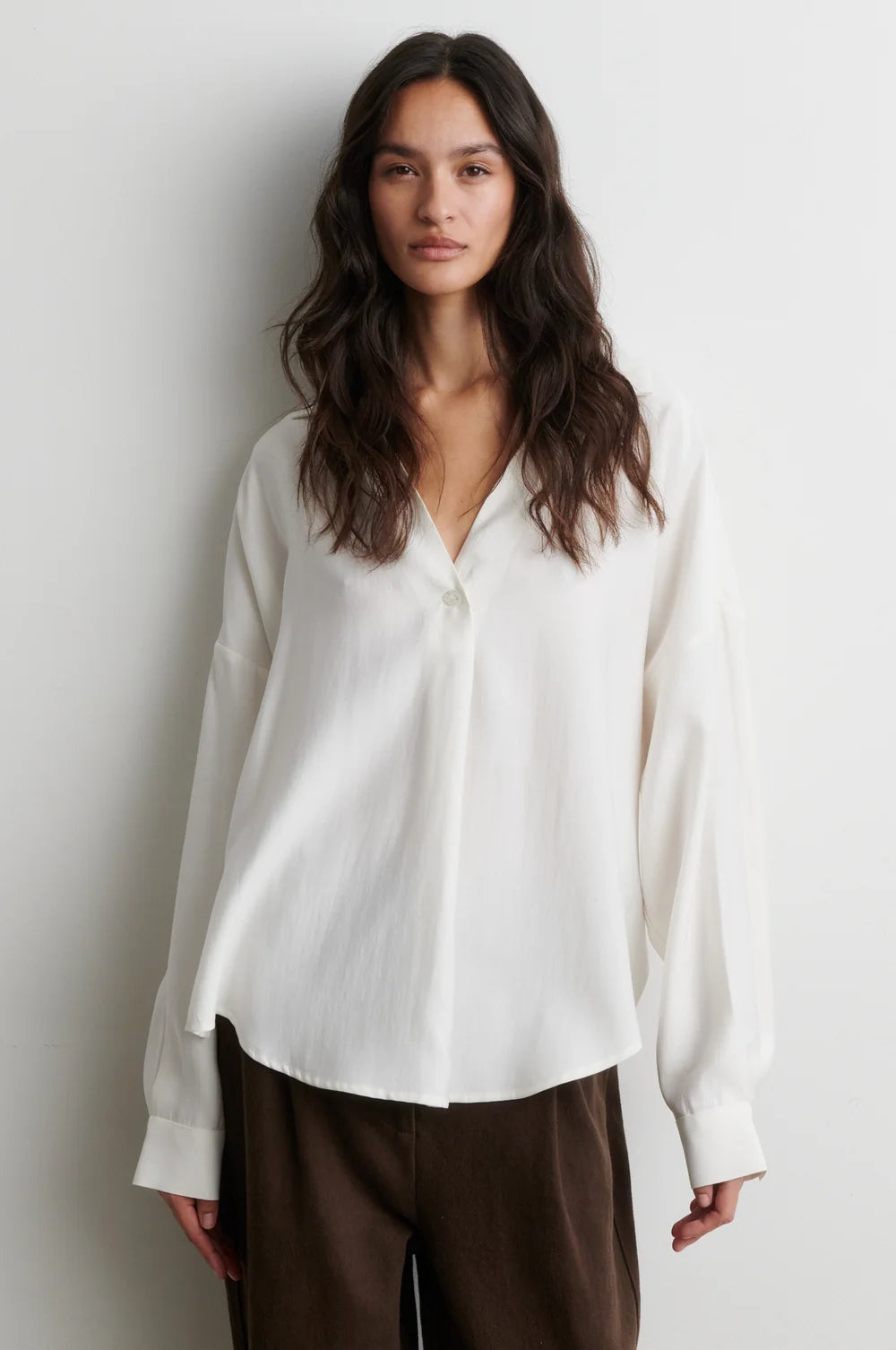 A woman modeling a white long-sleeve button-up blouse with a relaxed, slightly draped fit, paired with brown trousers, against a plain light gray wall.
