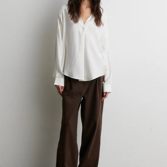 A woman stands against a plain light gray wall wearing a loose white long-sleeve blouse and dark brown wide-leg trousers.