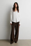 A woman stands against a plain light gray wall wearing a loose white long-sleeve blouse and dark brown wide-leg trousers.