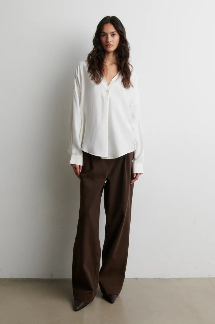 A woman stands against a plain light gray wall wearing a loose white long-sleeve blouse and dark brown wide-leg trousers.