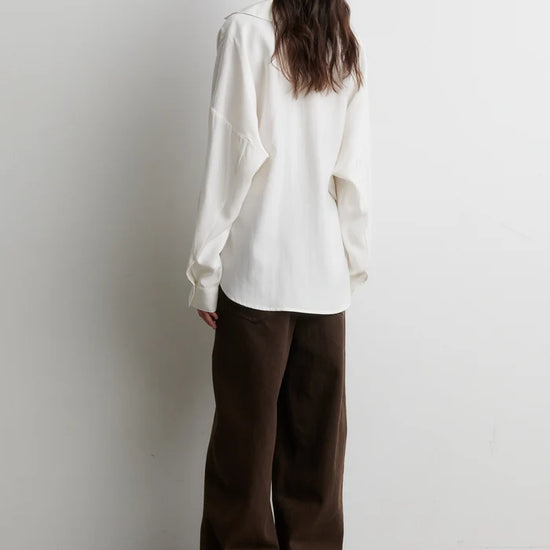 A woman stands with her back to the camera wearing an oversized white long-sleeve shirt and dark brown wide-leg trousers, set against a plain white wall and light concrete floor.