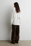A woman stands with her back to the camera wearing an oversized white long-sleeve shirt and dark brown wide-leg trousers, set against a plain white wall and light concrete floor.