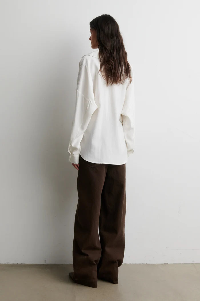 A woman stands with her back to the camera wearing an oversized white long-sleeve shirt and dark brown wide-leg trousers, set against a plain white wall and light concrete floor.
