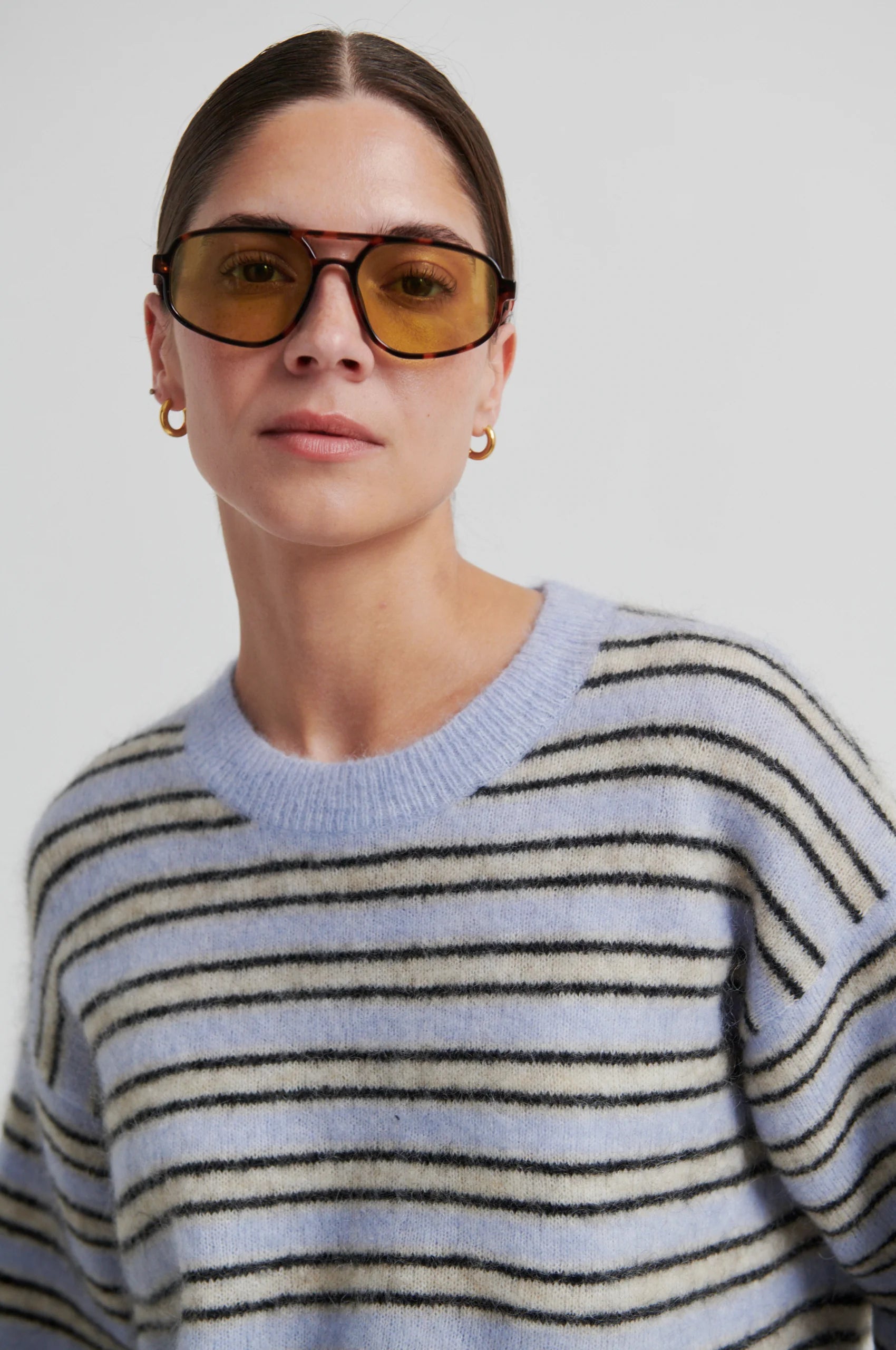 Person wearing a light blue and beige striped sweater with yellow-tinted sunglasses against a white background from Second Female