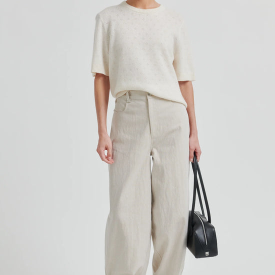 Woman wearing a white top and beige pants holding a black handbag on a light gray background from second female