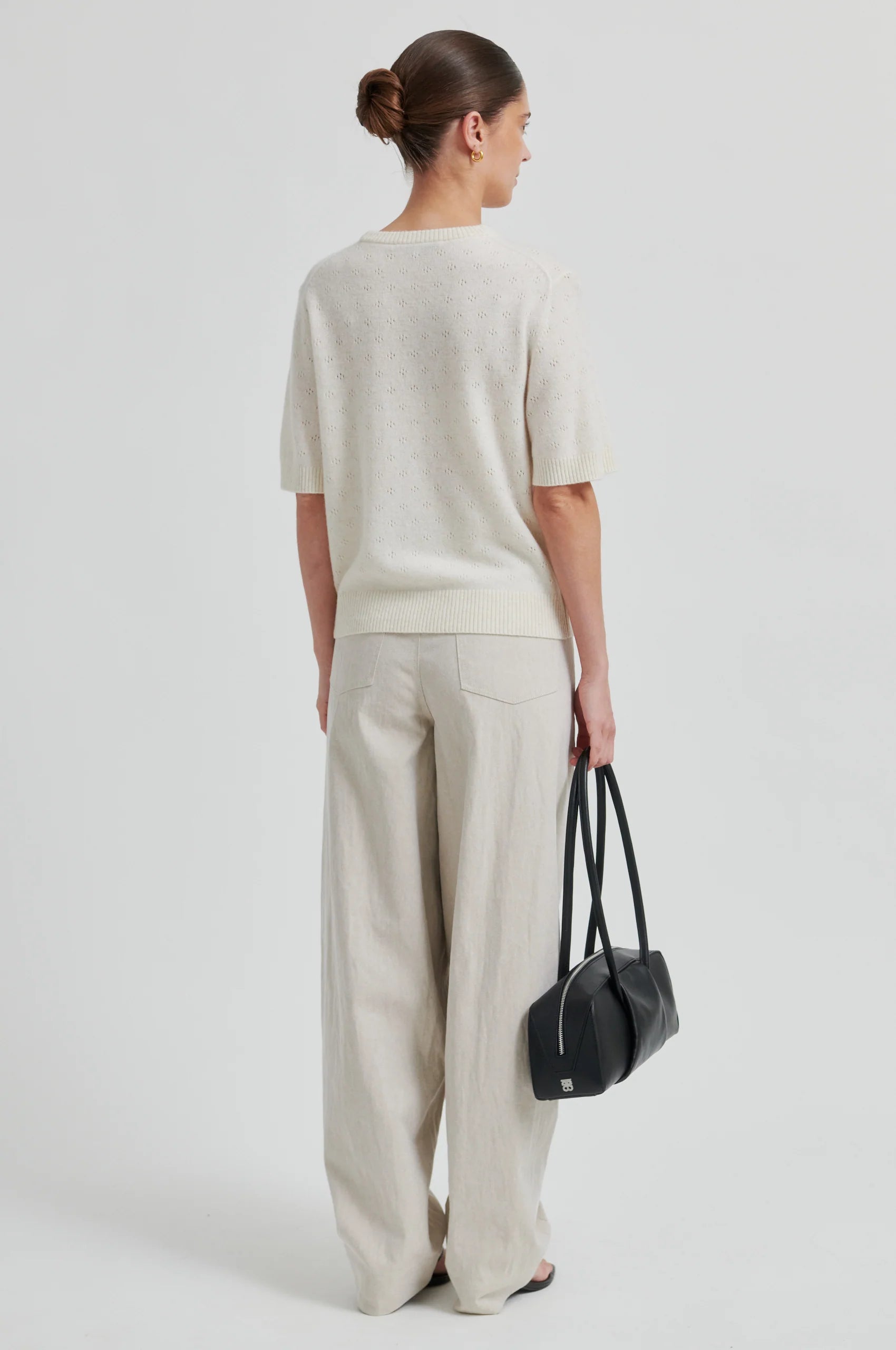 Woman wearing a white top and beige pants, holding a black handbag on a light gray background from second female